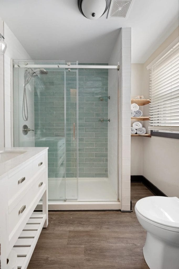 Modern bathroom with a glass-enclosed shower featuring light blue subway tiles, a white vanity with drawers, a white toilet, wood flooring, shelves with rolled towels, and a window with white blinds.
