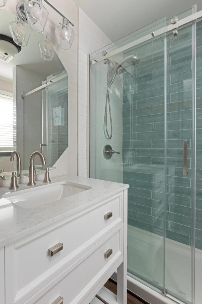 Modern bathroom with a white vanity, dual sinks, and chrome fixtures next to a glass-enclosed shower featuring light blue subway tiles and a mounted showerhead.