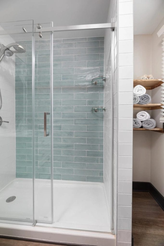 A modern shower with glass sliding doors, light blue subway tile on the back wall, chrome fixtures, and shelves holding neatly rolled white and gray towels beside it.