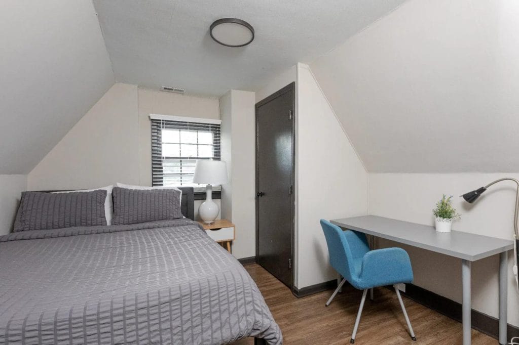 Small, tidy bedroom with a gray bed, nightstand with lamp, window with blinds, gray door, and a minimalist desk setup featuring a blue chair, desk lamp, and a small potted plant.