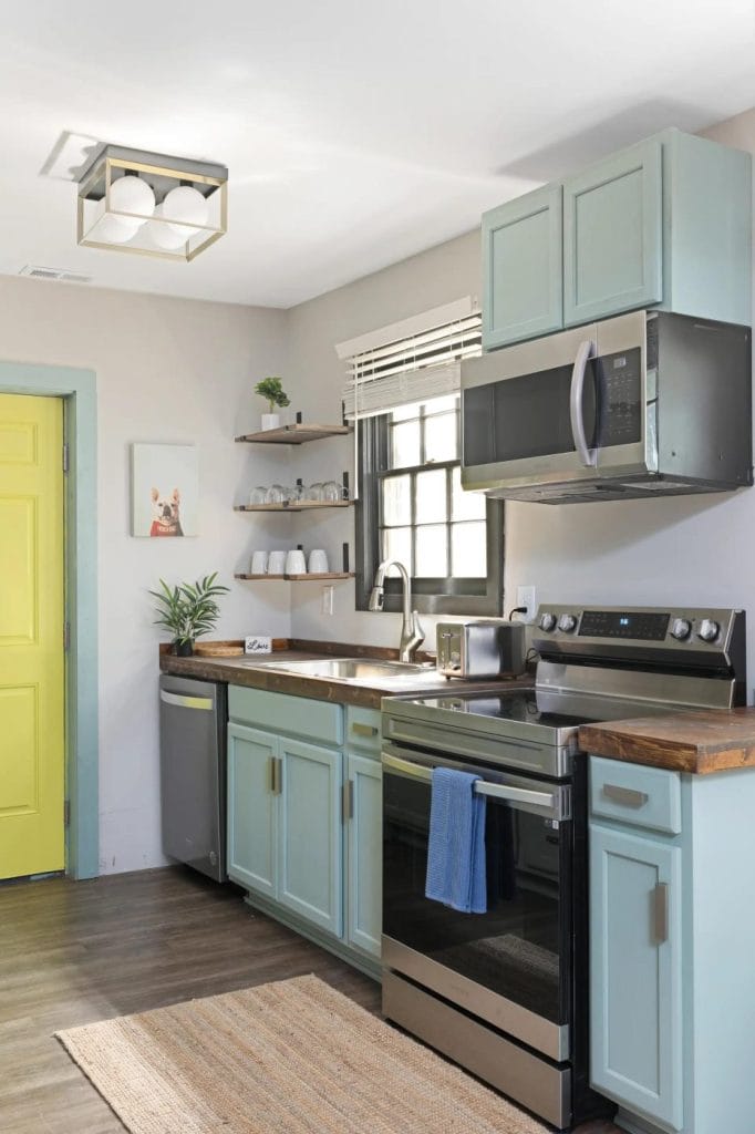 A modern kitchen with pastel blue cabinets, wood countertops, stainless steel appliances, open shelves with dishes, a yellow door, a window above the sink, and a beige rug on a dark wood floor.