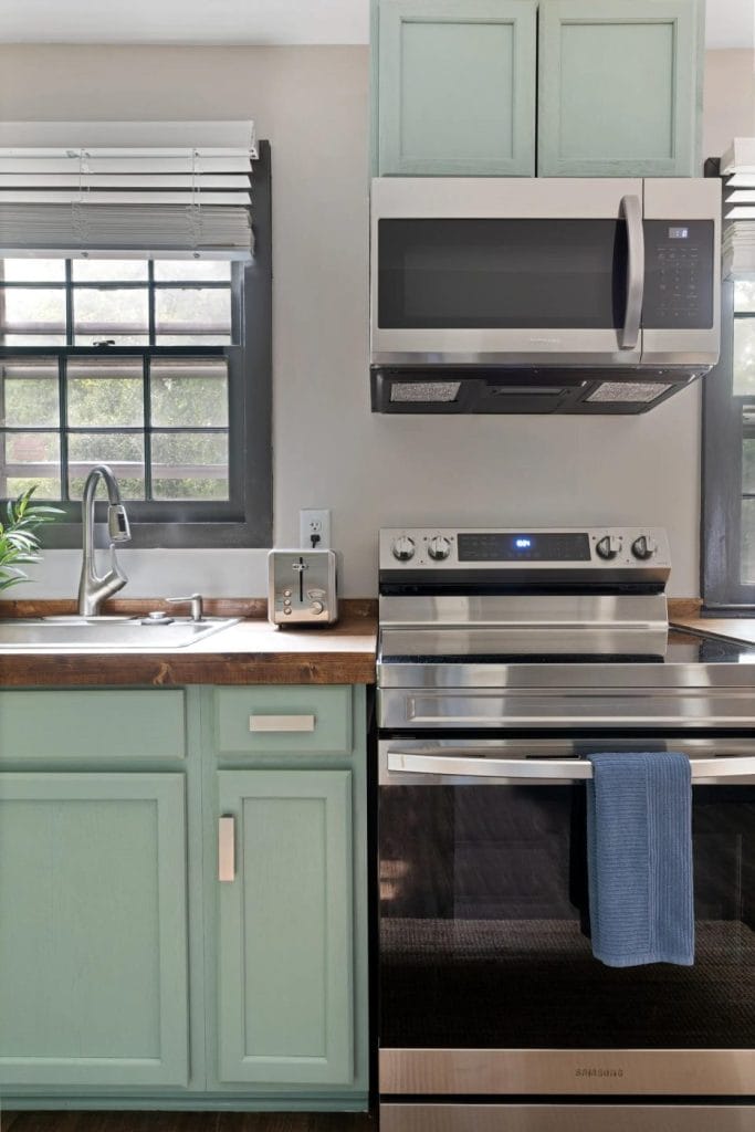 A modern kitchen with mint green cabinets, stainless steel oven and microwave, a sink with a window above it, a toaster, and a blue towel hanging from the oven handle.
