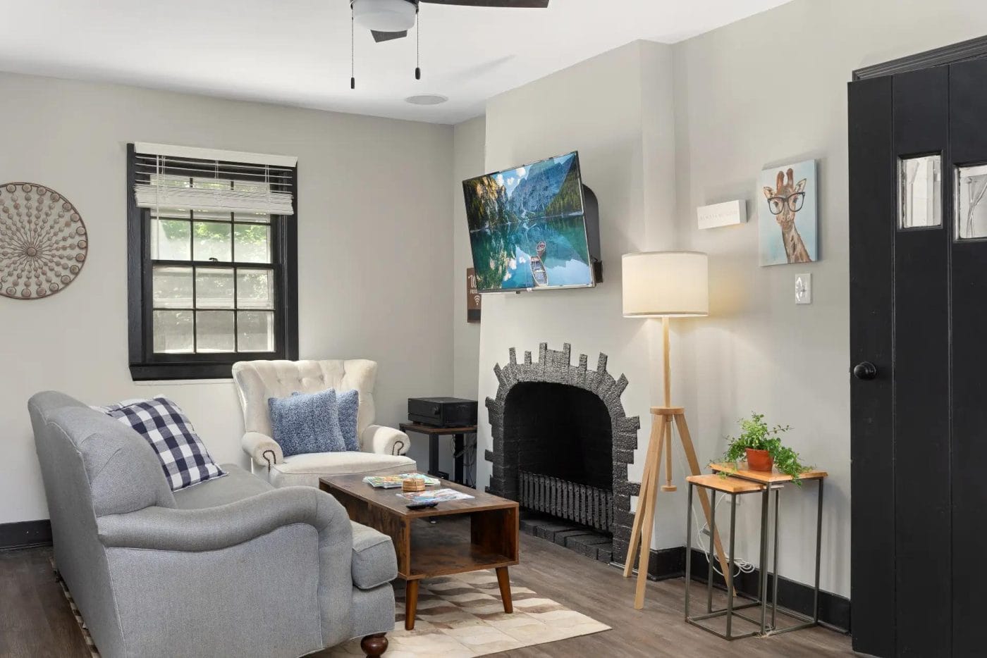 A cozy living room with a gray sofa, white armchair, wooden coffee table, TV mounted above a black fireplace, floor lamp, small side table with plant, and wall art. There’s a window with blinds and a dark door.