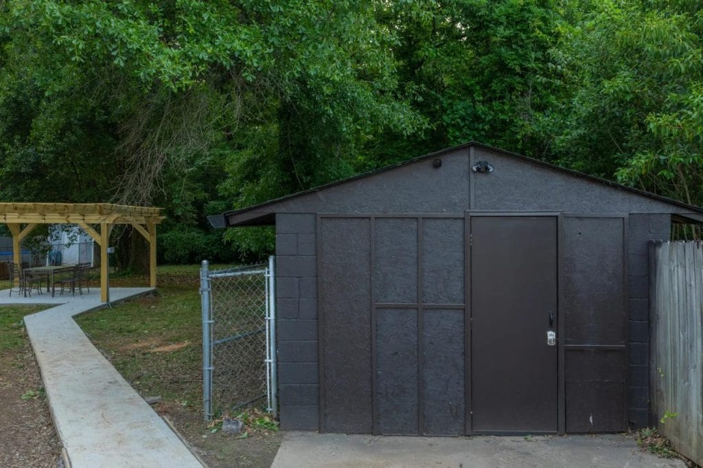 A small, dark-colored shed with a metal door sits next to a chain-link fence and wooden fence. In the background, there is a wooden gazebo with outdoor seating, surrounded by lush green trees.