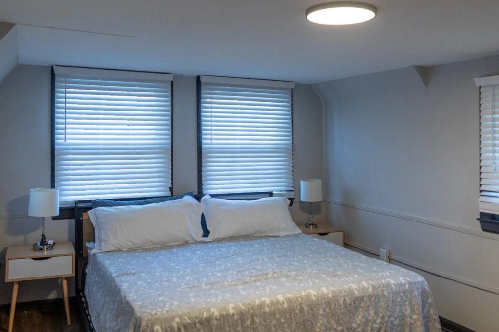 A neatly made bed with a patterned light gray comforter is centered between two windows with closed blinds. Two white lamps sit on modern nightstands on either side of the bed in a softly lit, minimalist bedroom.