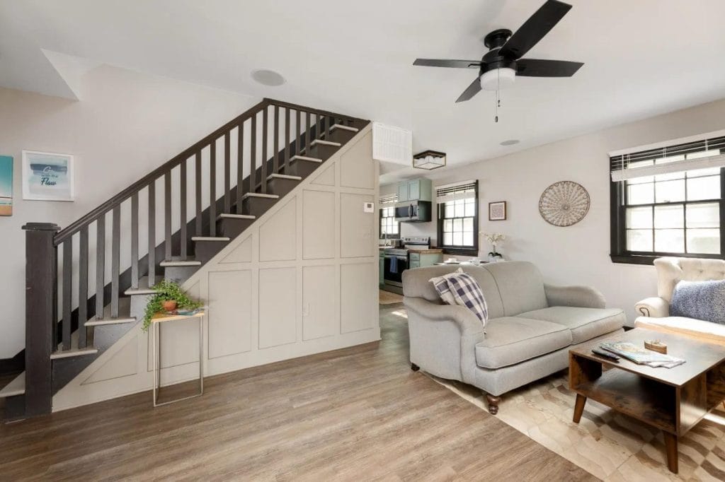 A cozy living room with a gray sofa, wooden coffee table, armchair, and a staircase with dark railings. The space has wood flooring, neutral walls, a ceiling fan, and large windows letting in natural light.
