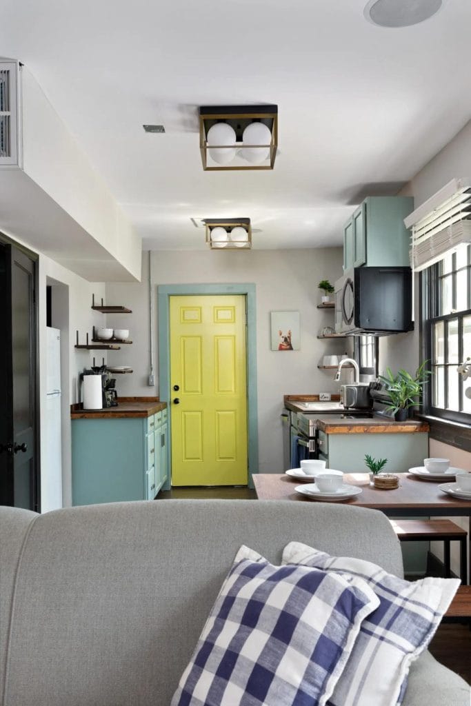 A cozy kitchen with blue cabinets, wooden countertops, open shelving, and a bright yellow door. A dining table is set, and a gray couch with a checkered pillow is in the foreground. Natural light fills the space.