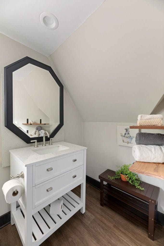 A small bathroom with a white vanity, octagonal mirror, stacked towels on a wooden bench, potted plant, and light walls under a sloped ceiling. The floor is dark wood.