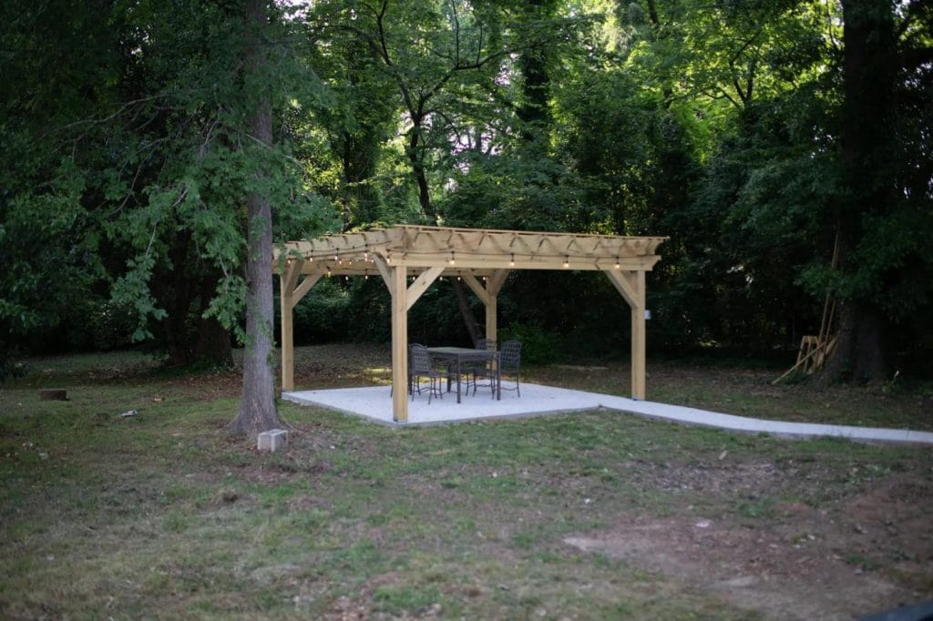 A wooden pergola stands on a concrete pad in a grassy, wooded area, with a table and several chairs underneath. The surrounding trees create a lush, green backdrop in the outdoor setting.