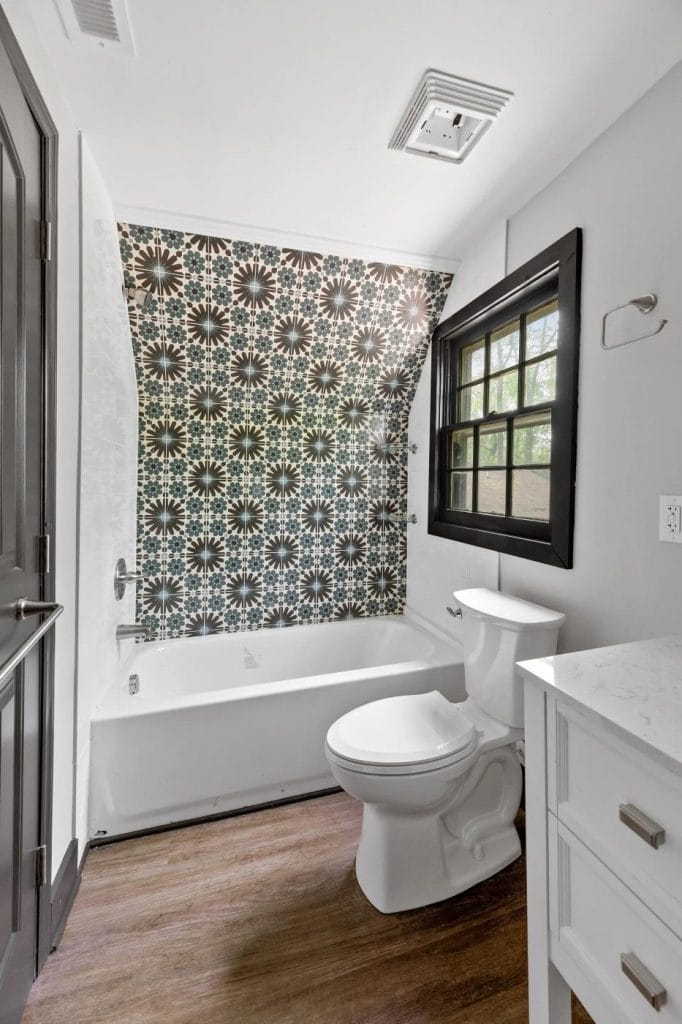 A modern bathroom with a patterned black and white tile accent wall above a white bathtub, a white toilet, a white vanity with marble countertop, wood-look flooring, and a black-framed window.