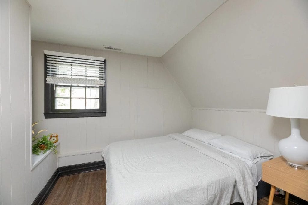 A small bedroom with a bed covered in white linens, a wooden nightstand with a white lamp, a window with blinds, and a plant on the windowsill. The walls are light-colored and the flooring is dark wood.
