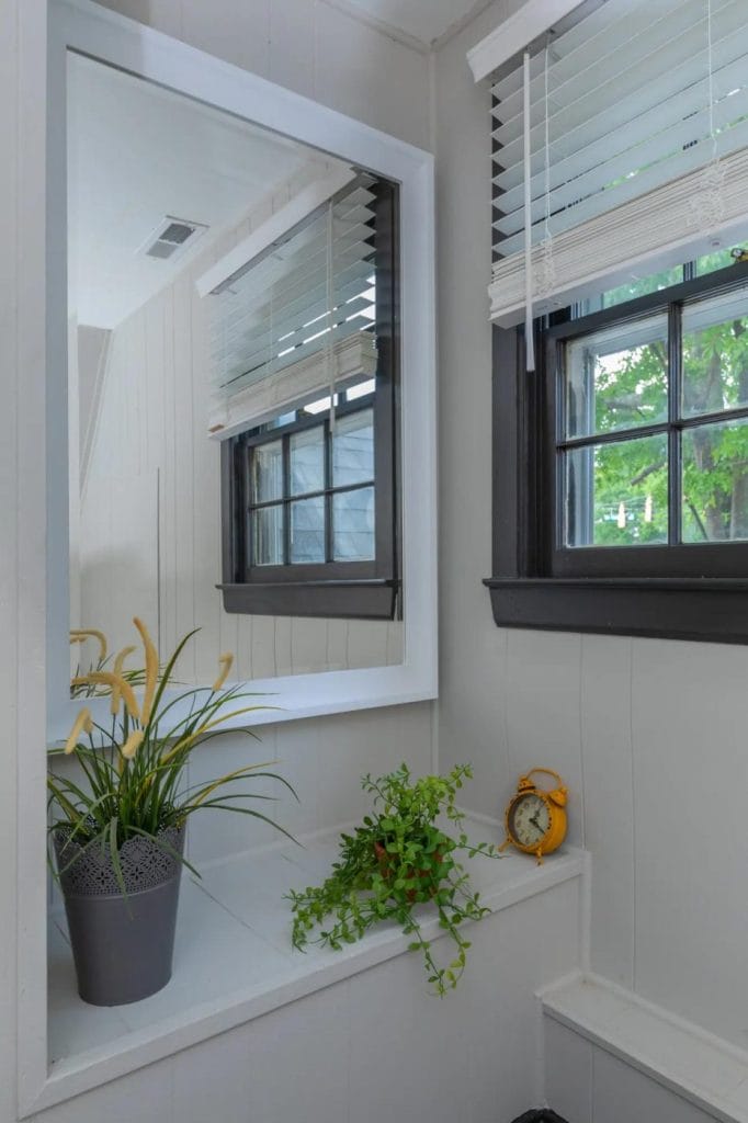 A corner of a room with a large mirror, two windows with white blinds, a potted plant, a small green leafy plant, and a yellow alarm clock on a white ledge. Green trees are visible outside.