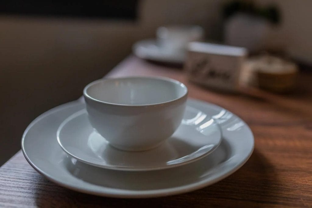A white ceramic cup sits on a matching saucer and dinner plate, neatly stacked on a wooden table. The background is softly blurred, showing more tableware and a small decorative sign.