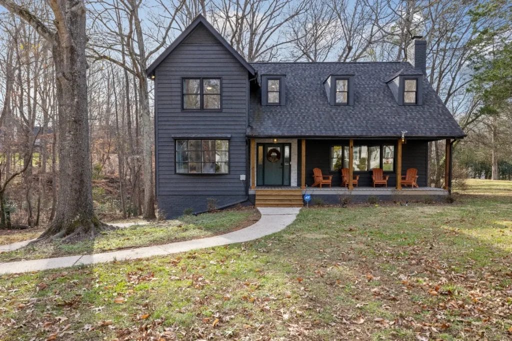 A dark gray two-story house with a covered front porch, several wooden chairs, and large windows. Leafy trees surround the lawn, and a curved concrete path leads to the front door. The ground is covered with fallen leaves.