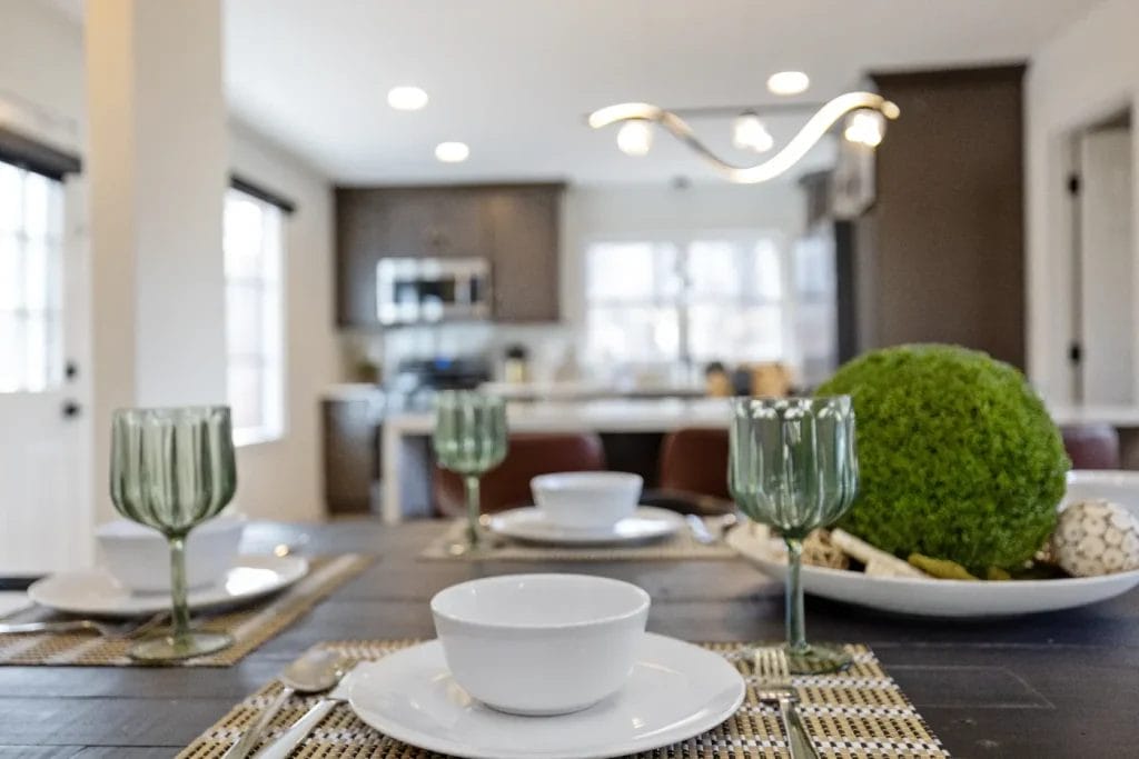 A set dining table with white bowls, green glass goblets, and placemats; a decorative centerpiece with moss balls; a bright, modern kitchen with stainless steel appliances is visible in the background.