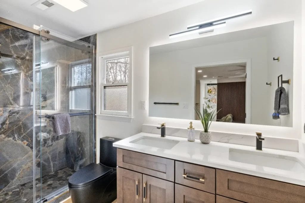 Modern bathroom with double-sink vanity, white countertop, large mirror, dark wood cabinets, black fixtures, black toilet, and a glass shower with dark marble tiles. Orchid plant and towel add decor.