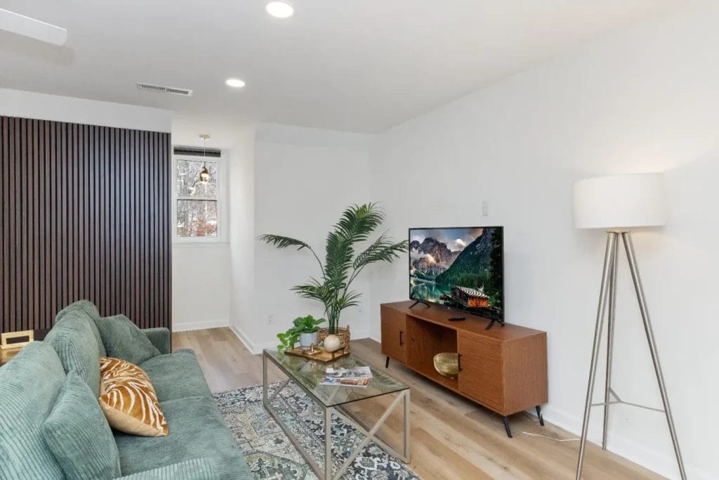 A modern living room with a green sofa, patterned rug, glass coffee table, potted plant, wooden TV stand with a flat-screen TV, and a tripod floor lamp on light wood flooring and white walls.