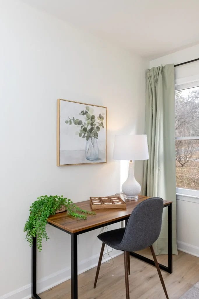 A small modern desk with a gray chair, a table lamp, a potted plant, and a decorative tray sits by a window with light green curtains. A framed painting of a vase with leaves hangs on the white wall above.
