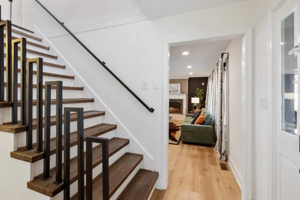 A modern staircase with wooden steps and black railings leads to a bright hallway. Through an open doorway, a living room with a green sofa, fireplace, and patterned curtains is visible.