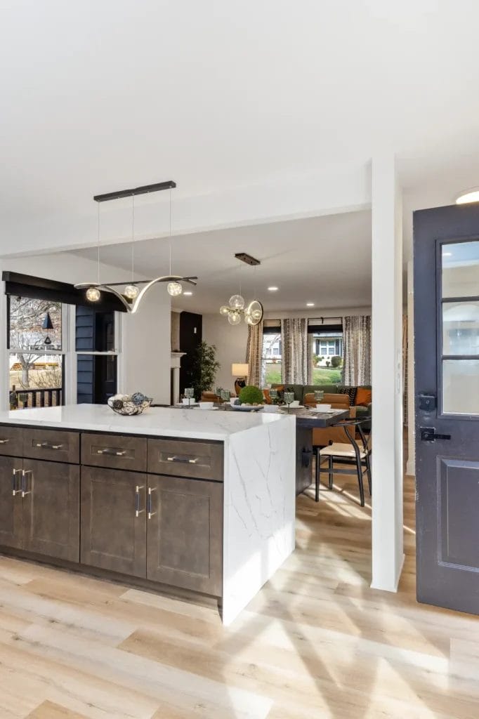 Modern kitchen with a marble island, dark cabinets, and gold pendant lights. Open floor plan shows a glimpse of a dining area and living room with large windows and light wood flooring throughout.