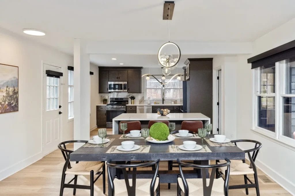 A modern dining area with a black table set for six, featuring white dishes and green glasses. Behind it is a kitchen with dark cabinets, stainless steel appliances, and an island with three brown stools. Large windows let in natural light.