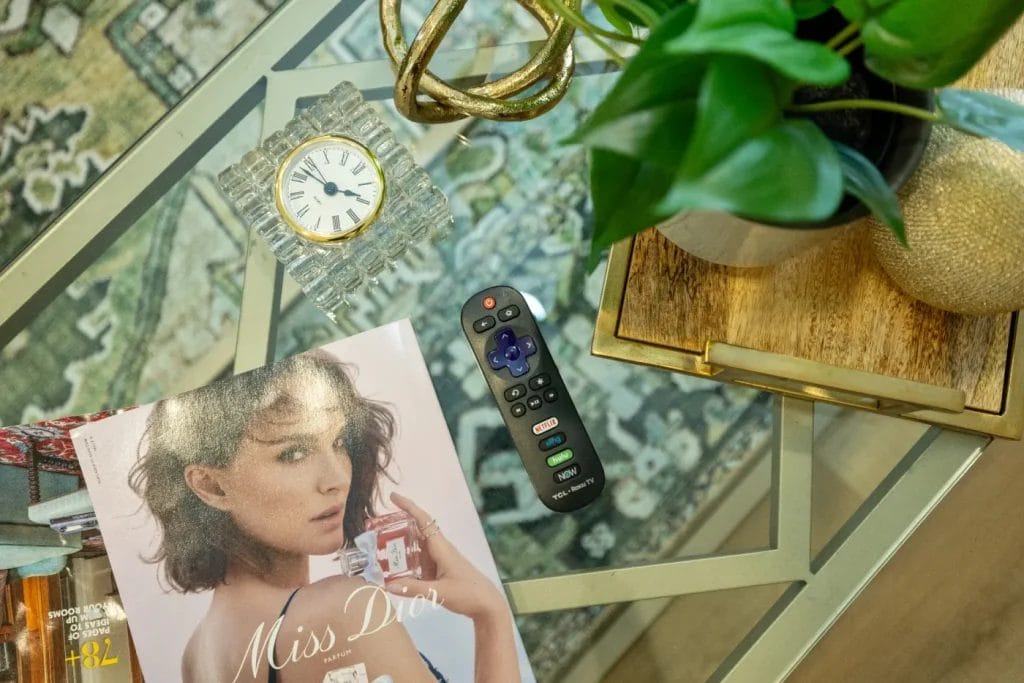A glass coffee table with a magazine featuring a woman holding a perfume, a TV remote, a decorative clock, and a potted plant on a wooden tray, all set atop a patterned rug.