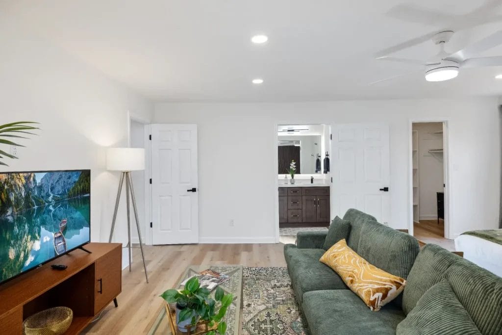 A modern living room with a green sofa, yellow pillow, patterned rug, TV on a wooden stand, floor lamp, and view into a bathroom with double sinks through open doors. Light wood flooring and white walls throughout.