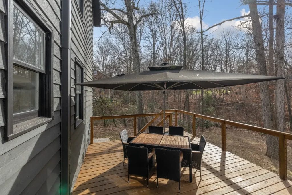 A wooden deck with a square table, six black chairs, and a large umbrella is attached to a gray house. The deck overlooks a wooded area with leafless trees under a partly cloudy sky.