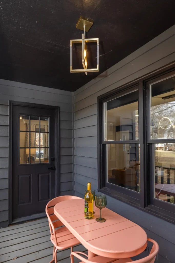 A small porch with dark siding features a peach-colored bistro table and two matching chairs. On the table are a green bottle, a wine glass, and a candle. A modern overhead light hangs from the black ceiling.