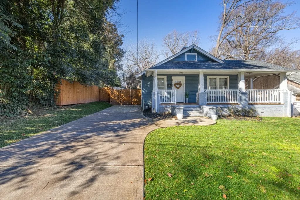 A small blue house with a front porch, white railings, and a heart-shaped wreath on the door. A long driveway curves past a green lawn, and a wooden fence lines the property under tall trees on a sunny day.