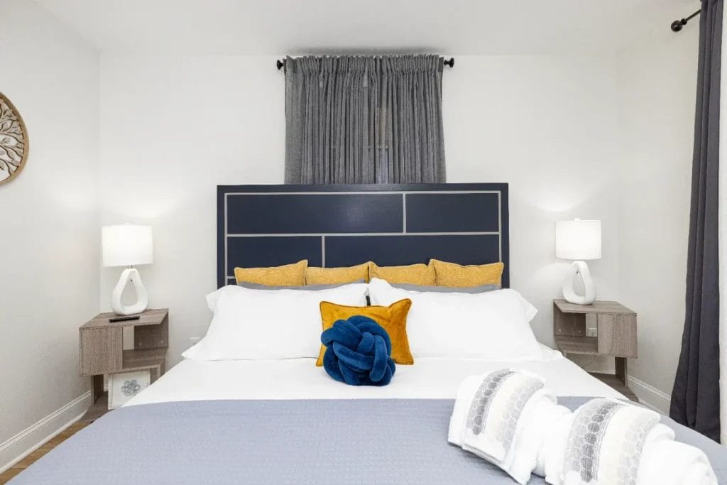 A modern bedroom with a large bed, white bedding, yellow accent pillows, and a blue towel folded on top. There are two bedside tables with lamps and a dark gray curtain behind the headboard. The room has a clean, minimal style.