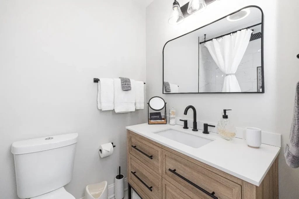 Modern bathroom with a wooden vanity, white countertop, and black fixtures. Above the sink is a large rectangular mirror with lights. Towels hang on the wall, and a white shower curtain is visible in the background.