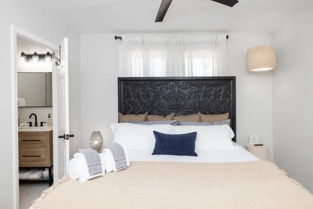 A neatly made bed with white and beige bedding, a dark carved headboard, and a navy accent pillow. Towels are folded at the foot of the bed. A bathroom with a sink is visible through an open door on the left.