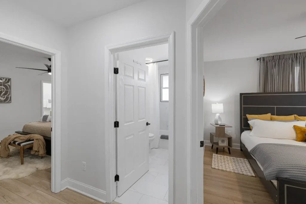 A bright hallway with white walls and wood floors leads to a bathroom with white tile and two bedrooms, both with beds, nightstands, and neutral-colored decor. Natural light fills each room.