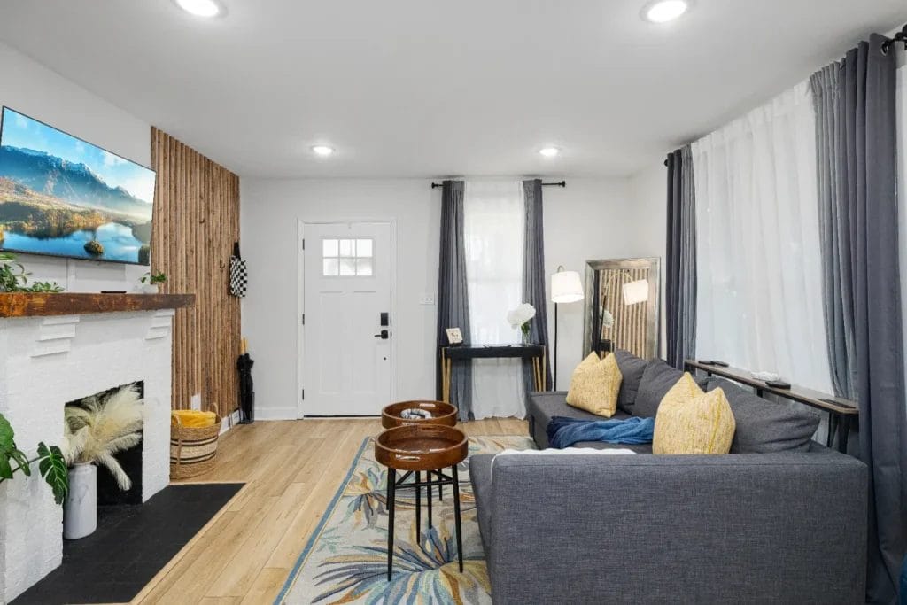 Modern living room with a gray sofa, yellow pillows, two round wooden tables, a white fireplace with a mounted TV, wooden accent wall, and large windows with sheer and dark curtains. Light wood floor and a patterned rug.