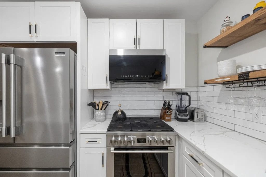 Modern kitchen with white cabinets, stainless steel refrigerator and stove, white subway tile backsplash, marble countertops, and wooden shelves holding dishes and glassware. Appliances and utensils are neatly arranged on the counter.