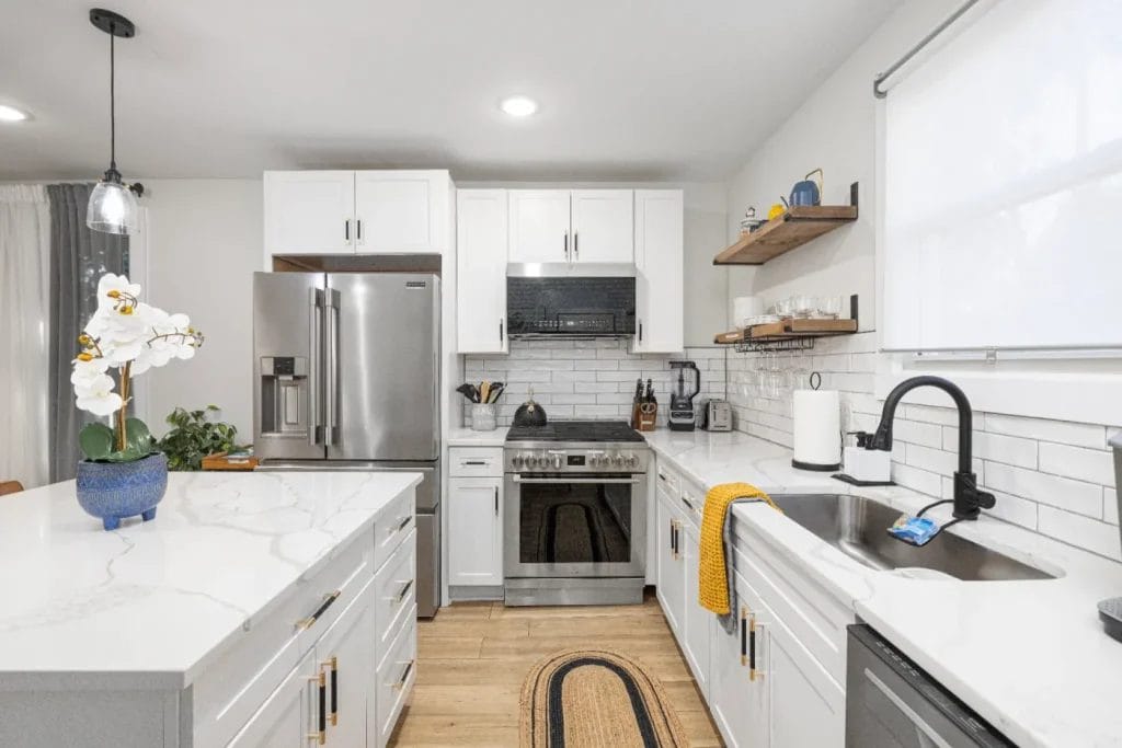 Modern kitchen with white cabinets, marble countertops, stainless steel appliances, a gas stove, open wooden shelves with dishes, a sink by the window, and a blue pot with a white orchid on the island.
