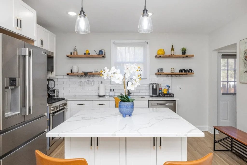 Modern kitchen with white cabinets, stainless steel appliances, open wooden shelves, and a large white marble island. Two pendant lights hang above the island, which has a blue vase with white flowers.