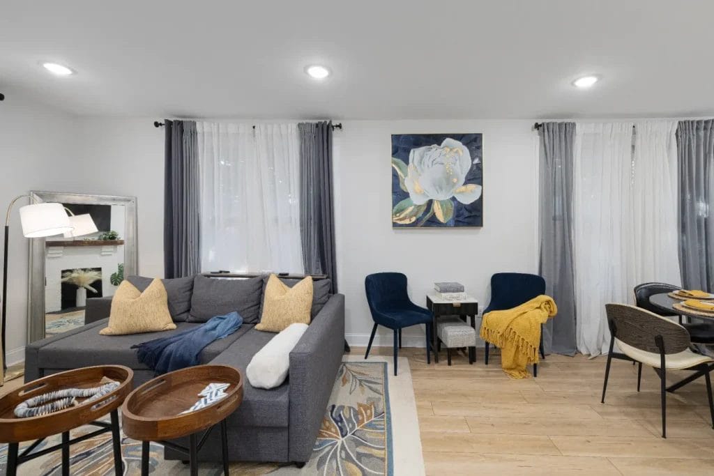 Modern living room with a gray sectional sofa, yellow pillows, two blue chairs, a floral wall painting, dining table, and floor-length curtains. Light wood floors and accent decor create a cozy, inviting atmosphere.