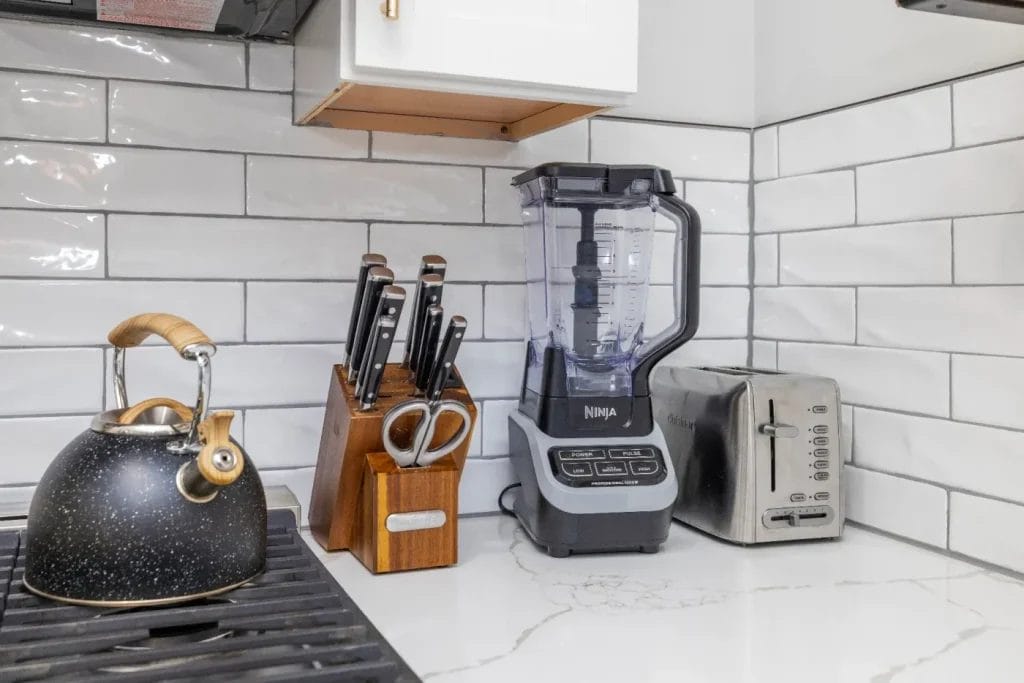 A black kettle on a stovetop sits next to a wooden knife block, a Ninja blender, and a stainless steel toaster on a white marble countertop with white subway tile backsplash.