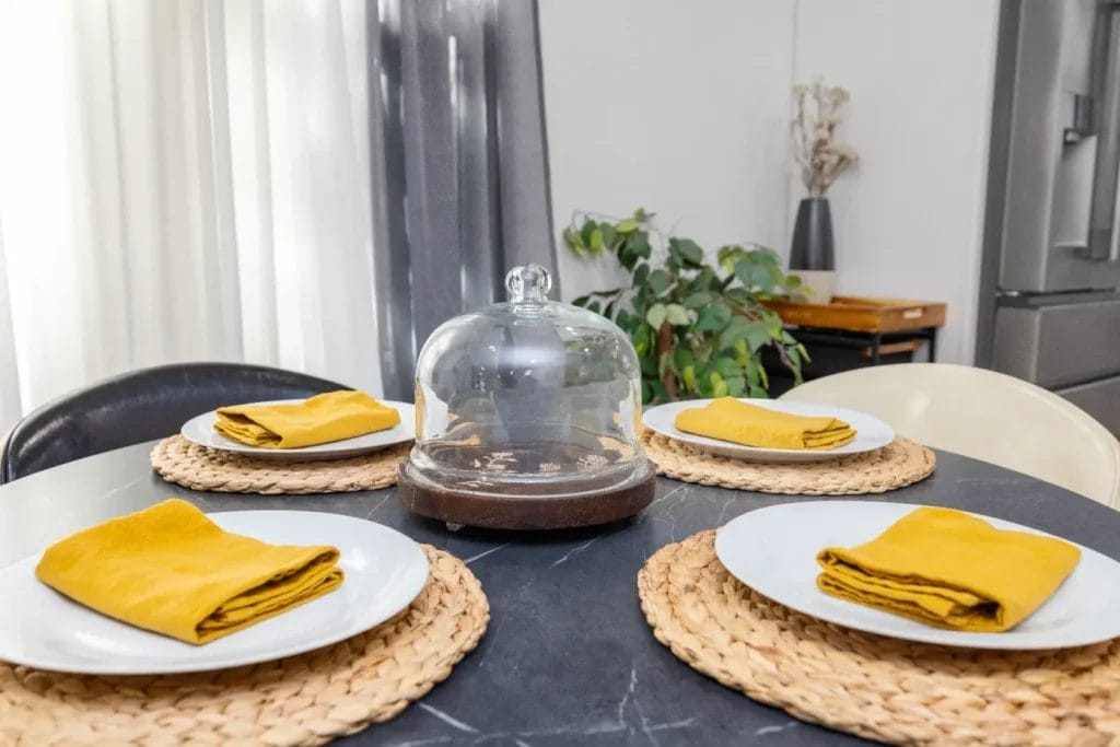 A round dining table set for four with white plates, yellow napkins, woven placemats, and a glass cloche centerpiece. A plant and decorative items are in the background near a window with sheer curtains.