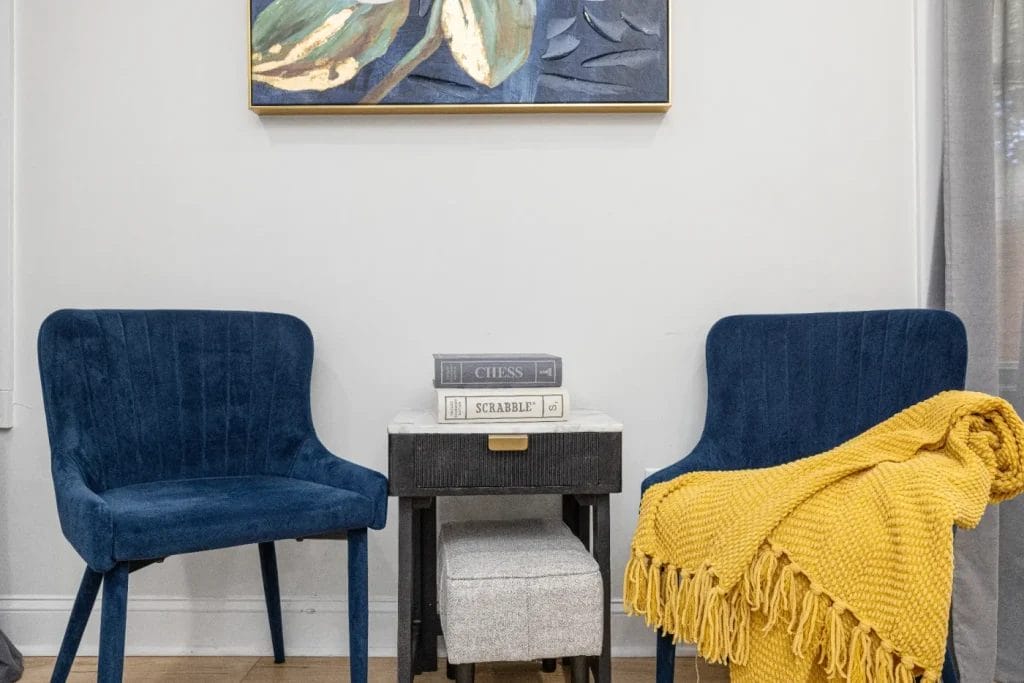 Two blue velvet chairs sit on either side of a small black table with books stacked on top. A yellow blanket is draped over one chair, and a colorful painting hangs on the wall above them.