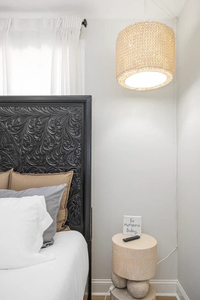A cozy bedroom corner with a carved dark headboard, neutral pillows, a round wooden nightstand, a small sign reading "be awesome today," and a woven pendant light hanging above. White curtains cover the window.