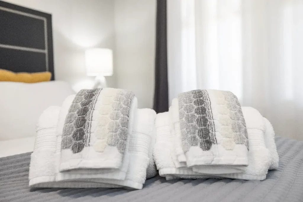 Two neatly folded stacks of white towels with gray patterned hand towels on top are placed on a bed with a gray cover in a softly lit, modern bedroom. A bedside lamp glows in the background.