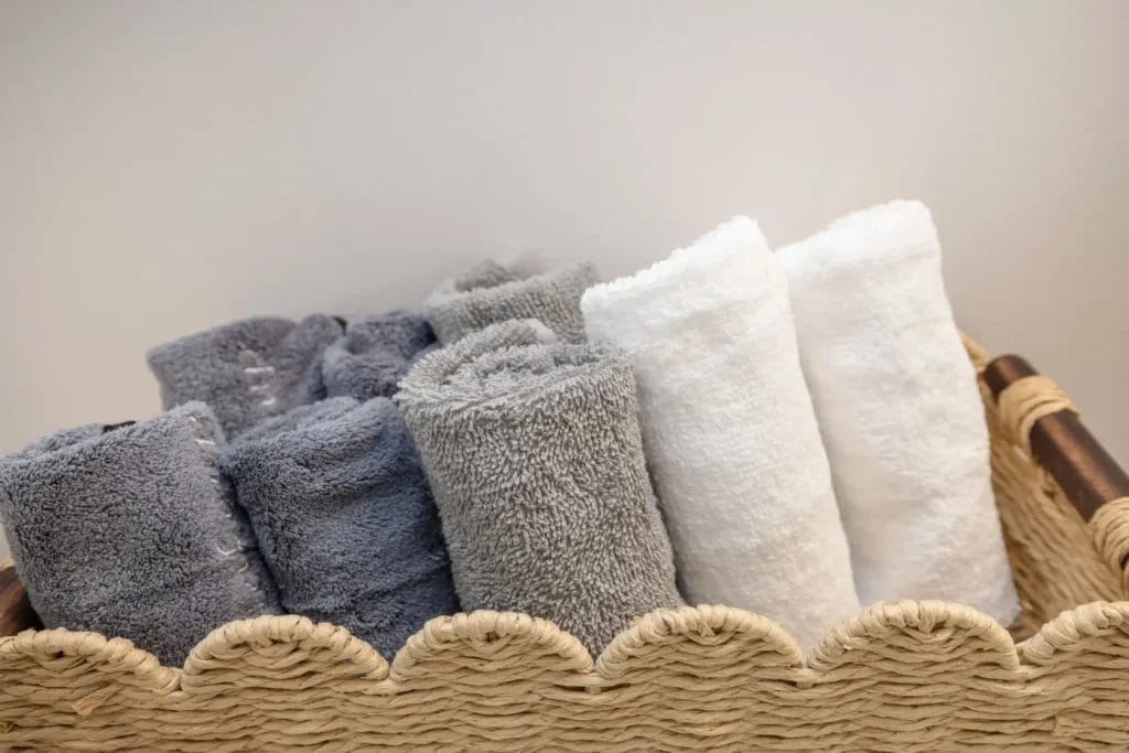 A woven basket holds several neatly rolled towels, with gray towels on the left and white towels on the right, against a plain light-colored background.