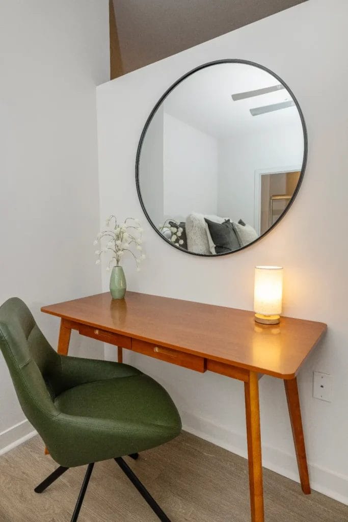 A minimalist workspace with a wooden desk, green swivel chair, round wall mirror, small lamp, and a vase with white flowers against a white wall and wood flooring.