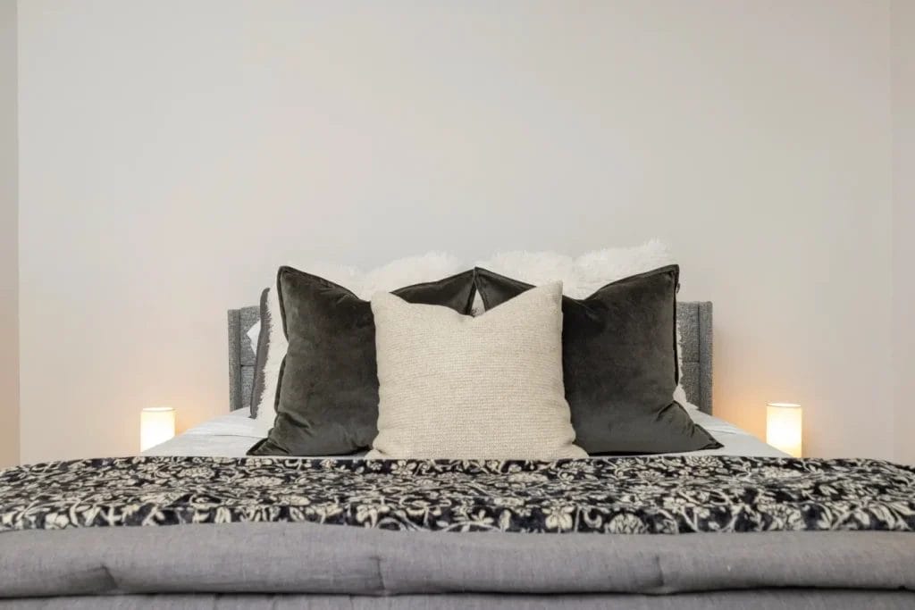 A neatly made bed with three large throw pillows—two dark and one light—in front of a gray upholstered headboard. A patterned blanket is folded at the foot, with two small lit lamps on each side.