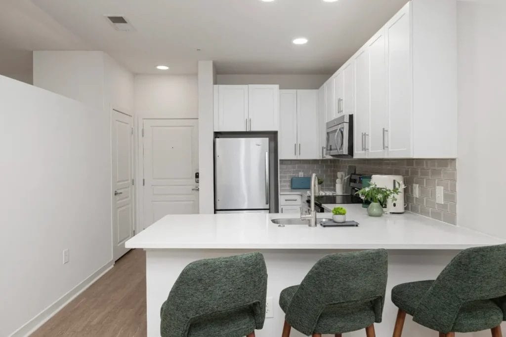 Modern kitchen with white cabinets, stainless steel appliances, gray tile backsplash, and a large white island with a sink. Three green upholstered barstools are placed at the island. A plant and small decor items sit on the counter.