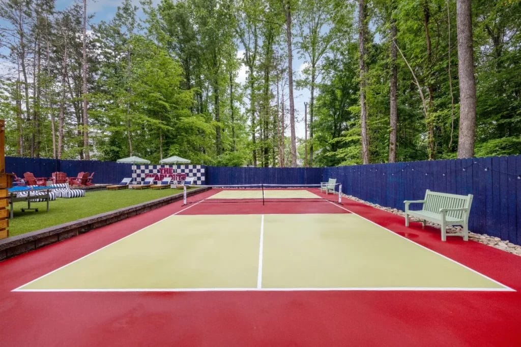 A small outdoor pickleball court with red and yellow surfaces, surrounded by a blue fence and tall trees. Benches and Adirondack chairs are on the sidelines, with a checkered sign in the background.