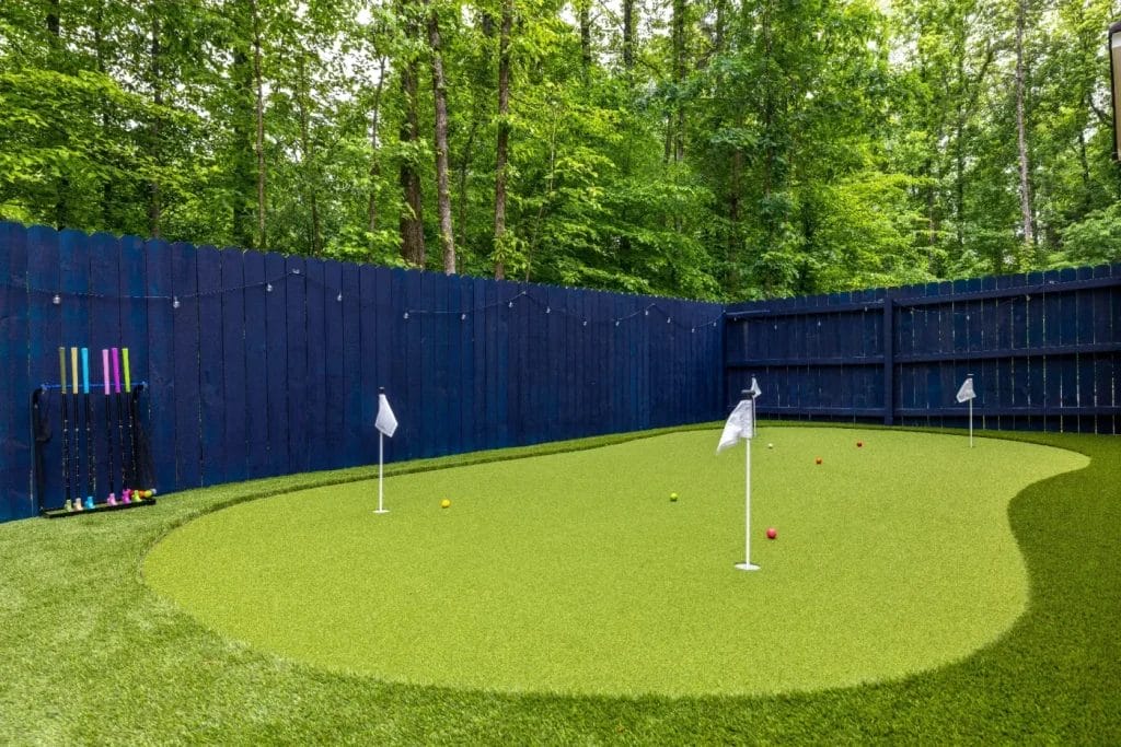 A backyard putting green with three flags, surrounded by a tall navy blue wooden fence and trees. Four colorful mini golf putters and balls are arranged on the left side near the fence.
